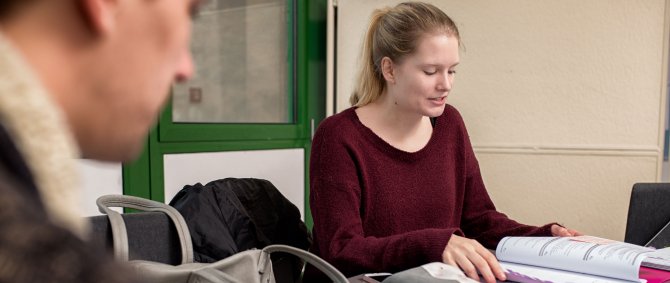 Eine Studentin blättert in vor ihr auf einem Tisch liegenden Unterlagen. Links im Bild ist angeschnitten ein Student zu sehen, der mit ihr am Tisch sitzt.