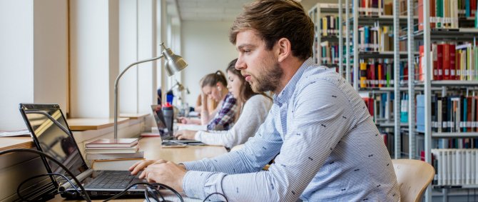 Eine Bibliothek. Rechts sind Bücherregale zu erkennen, links arbeiten mehrere Studierende an einem langen Tisch. Vorn im Bild ein kurzhaariger Mann im blauen Hemd, der an einem Laptop arbeitet.