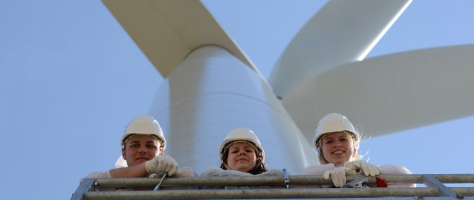 Drei junge Menschen schauen eher von oben in die Kamera, hinter ihnen ist blauer Himmel und eine Windenergieanlage zu erkennen.