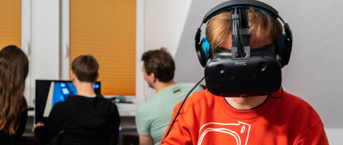 Im Vordergrund Person mit einer Virtual-Reality-Brille, im Hintergrund Personen vor einem Computermonitor.