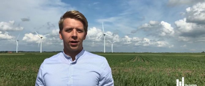 Junger Mann mit kurzen blonden Haaren und hellbaluem Hemd steht auf einer Wiese. Im Hintergrund Wolken am Himmel und Windräder.