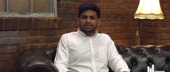 Junger Mann in weißem Hemd sitzt auf einem Sofa, hinter ihm ein Bilderrahmen und eine Stehlampe. Er spricht in die Kamera.