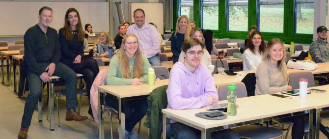 Seminarraum: Junge Menschen sitzen an Tischen und schauen in die Kamera. Auf einem Tisch sitzen vier Menschen: Die Lehrenden und die Unternehmensvertreter*innen.