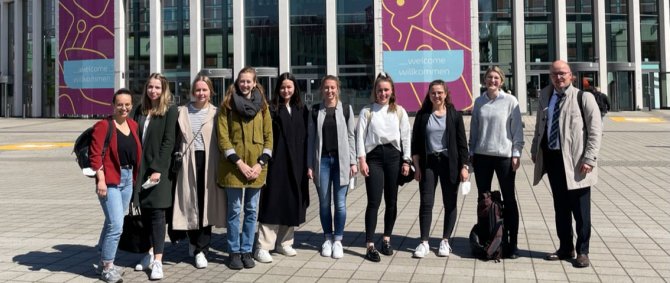 Eine Gruppe Studierender und ein Lehrender posieren vor der Messe Berlin. Am Gebäude hängt ein großes Plakat zur Messe.