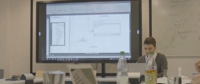 Junger Mann sitzt an Besprechungstisch, hinter ihm ein großer Bildschirm, auf dem Tisch Papier und Getränke.