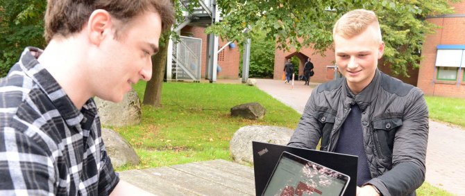 Zwei junge Männer sitzen draußen an einem Tisch und schauen auf Laptops, die vor ihnen stehen.