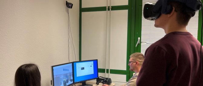 Ein Studierender mit VR-Brille und Controllern steht vor einem Arbeitstisch, an dem zwei weitere Studierende an ihren Computern arbeiten.