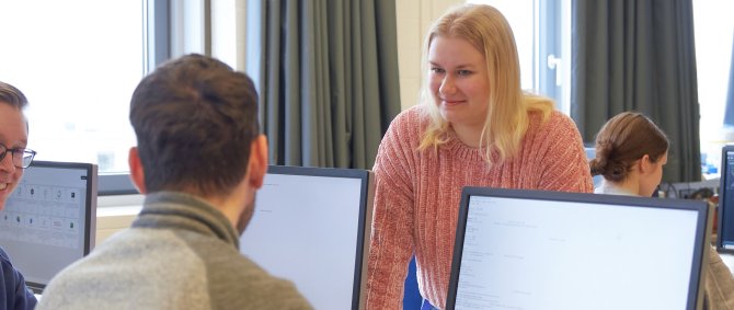 Studierende in einem Seminarraum. Zwei Studenten schauen auf Bildschirme, eine Studentin steht dahinter. Sie unterhalten sich.