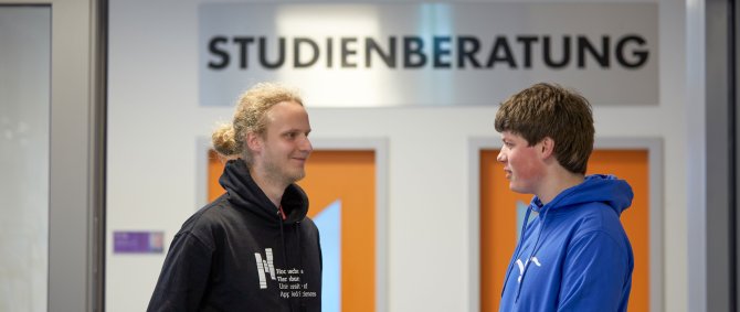 Zwei Studierende im Gespräch, hinter ihnen sind orange-farbene Türen und darüber ein Schild "Studienberatung" zu erkennen.