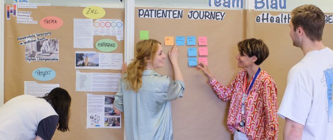 Studierende und Dozentin vor einer Pinnwand. Sie sind im Gespräch. Zu erkennen sind Wörter wie "eHealth" und "Patienten Journey".