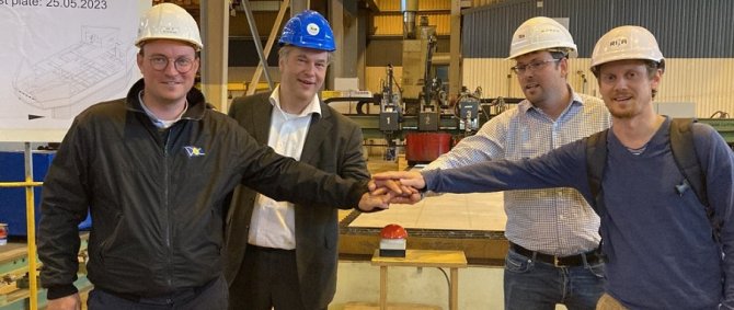 Vier Männer stehen in einer Werfthalle rund um einen großen roten Knopf. Sie haben die Hände übereinander auf den Knopf gelegt und lächeln in die Kamera. Alle vier tragen Bauschutz-Helme.