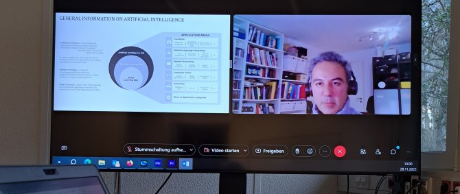 Auf einem großen Monitor ist ein Splitscreen mit einer Präsentation und einem Mann mittleren Alters zu sehen, der in die Kamera spricht. Die Präsentation hat den Titel &quot;AI Use Cases in Media&quot;. Im Vordergrund ein Tisch mit einem aufgeklappten Laptop, einem Schreibblock mit Notizen und einer Kaffetasse mit dem Logo der Hochschule Flensburg.