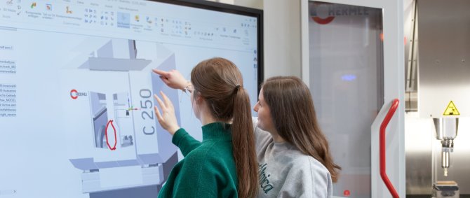 Zwei Studentinnen stehen an einem großen Monitor und zeigen auf die dort dargestellte Grafik mit verschiedenfarbigen eckigen Flächen und der Angabe C250. Ein Dozent steht links, ein Student links neben ihnen und blicken ebenfalls auf den Bildschirm.