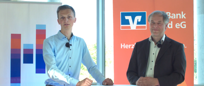 Ein Student und ein Dozent stehen an einem Stehtisch. Der Student spricht ernst blickend in die Kamera, der Dozent lächelt offen. Im Hintergrund zwei Aufsteller mit den Logos der Hochschule Flensburg und der VR Bank Nord eG.