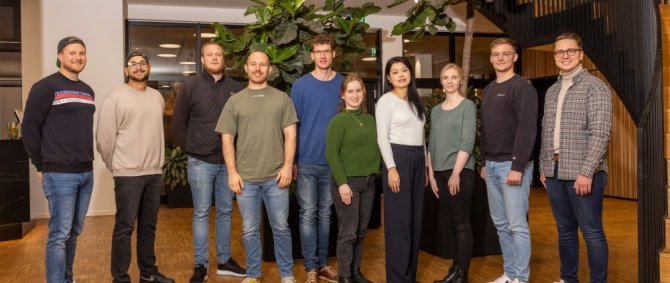 Zehn Studierende stehen in einer Reihe in einem Foyer mit vielen grünen Pflanzen und einer schwarzen Metalltreppe und lächeln in die Kamera.