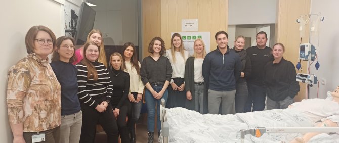 Eine Gruppe von jungen Menschen steht rund um ein Krankenhausbett, in dem eine Modellpuppe liegt. An der Wand hängt ein Monitor, neben dem Bett ein Tropf und an der Wand hinter den Studierenden hängt ein Poster mit Aufschrift in dänischer Sprache.