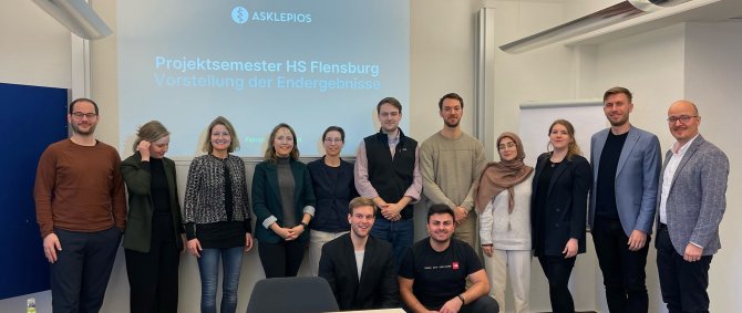 Eine Gruppe von überwiegend jungen Menschen steht in einer Reihe und lächelt in die Kamera, zwei junge Männer knien davor. Im Hintergrund ist die Projektion einer PPT-Folie zu sehen, auf der auf blauem Hintergrund das Logo der Asklepios-Gruppe zu sehen ist, darunter der Schriftzug: "Projektsemester HS Flensburg. Vorstellung der Endergebnisse".