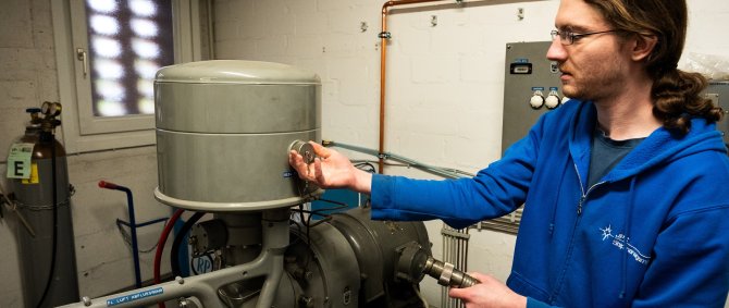 Ein junger Mann im blauen Kapuzenpulli steht an einem Flüssigluftgenerator, mit einer Hand an einem Verschluss und der anderen an einem Schlauch.