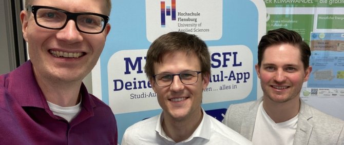 Drei Männer stehen nebeneinander und lächeln in die Kamera, im Hintergrund ein Aufsteller mit dem Logo der Hochschule Flensburg.