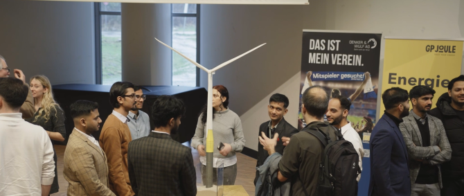 Eine Gruppe von Menschen steht um ein über menschengroßes Modell einer Windenergieanlage herum und untehält isich angeregt. Im Hintegrund sieht man Roll-Ups der Denker und Wulf AG und von GP Joule.