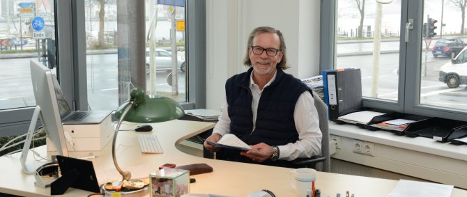 Ein älterer Mann mit schulterlangen grauen Haaren sitzt an einem Schreibtisch und lacht in die Kamera. Durch die Fenster im Hintergrund sieht man eine Straße und den Flensburger Hafen.