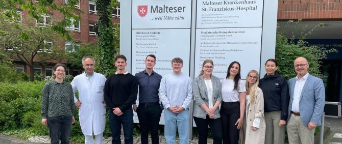 Eine Gruppe von Studierenden steht vor einem großen Backsteingebäude und zwei Wegweiser-Schildern, die es als das Malteser Krankenhaus St. Franziskus-Hospital ausweisen. Sie werden flankiert von einer Dame und einem Herrn im Arztkittel und einem Herrn im Anzug.