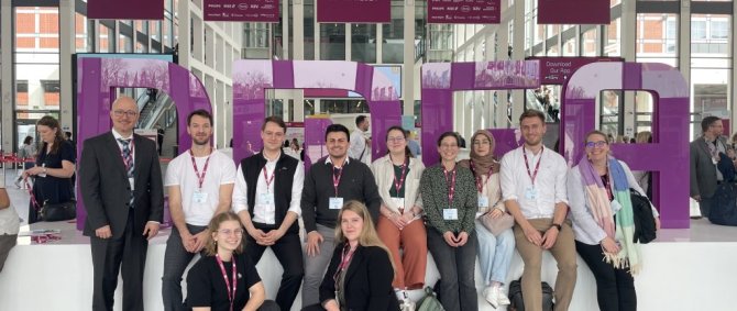 Eine Gruppe von Studierenden sitzt im Eingangsbereich einer Messehalle vor vier großen lilanen Buchstaben: DMEA.