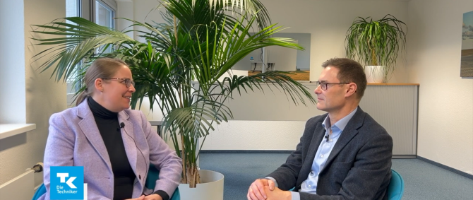 Eine Frau im lilanen Blazer und ein Mann im Anzug sitzen sich in einem großen Büroraum gegenüber und unterhalten sich. Im Hintergrund stehen zwei große Grünpflanzen.