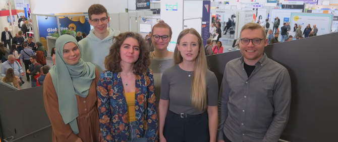 Eine Gruppe von jungen Menschen steht in einer Messehalle und lächelt in die Kamera.