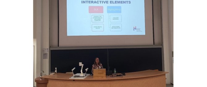 Eine junge Frau steht in einem Seminarraum an einem Pult. Hinter ihr an der Wand eine Projektion mit der Überschrift &quot;Interactive Elements&quot;.