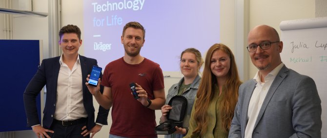 Drei Studierende und zwei Männer lächeln in die Kamera, im Hintergrund eine Projektion mit der Überschrift "Technology for Life - Dräger". Einer der Studierendne hält ein Smartphone und ein Messgerät, eine andere ein Schutzcase.