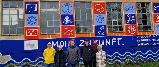 Eine Gruppe von Studierenden und ein Dozent steht vor einem niedrigen Gebäude, das bunt bemalt ist mit verschiedenen Logos und dem Slogan "Moin Zukunft".