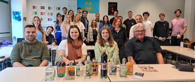 Zwei Männer und zwei Frauen sitzen in einem Seminarraum, vor ihnen ist eine Reihe von Glaschflaschen mit bunten Etiketten aufgebaut. Hinter ihnen steht eine große Gruppe von Studierenden.