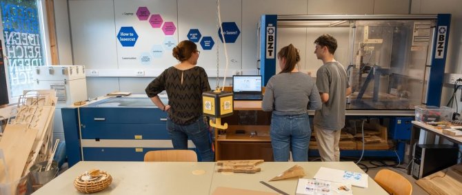 Studierende im ideenreich. Im Bildvordergrund ein Arbeitstisch mit Lineal, Notizbuch, Spanplatten, im Hintergrund ein Lasercutter.