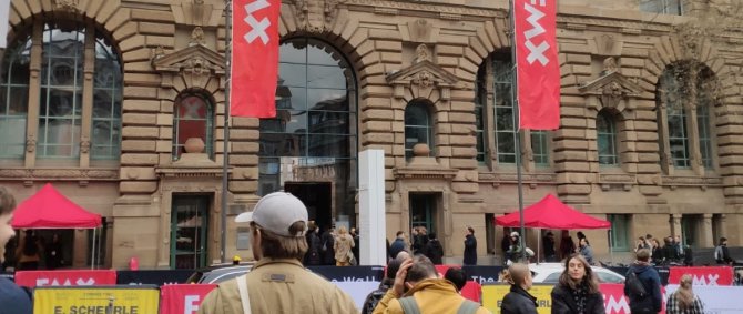 Eine Gruppe von jungen Menschen geht auf ein großes Sandsteingebäude zu, an dem rote Flaggen mit der Aufschrift "FMX" hängen.