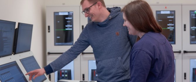 Ein Student und eine Studentin stehen in einem Raum mit vielen Monitoren, auf denen schiffsbetriebstechnische Anzeigen zu sehen sind.