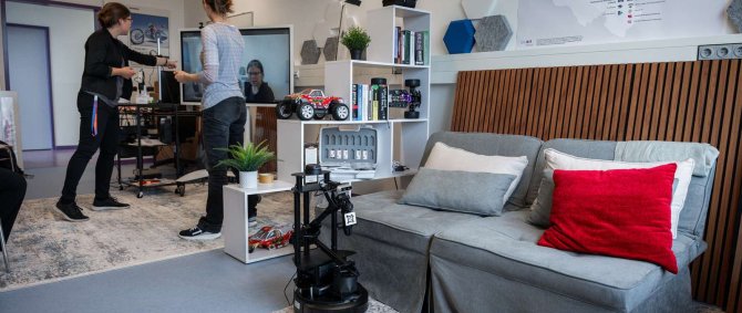 In einem Raum mit Sofa, Teppichen, Regal und Schreibtisch steht im Vordergrund ein Roboterarm auf dem Boden, im Hintergrund stehen zwei Frauen vor einem großen Monitor und deuten darauf.