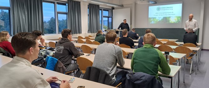 In einem Seminarraum sitzen Studierende in Tischreihen. Vorne stehen zwei Männer, hinter ihnen wird eine Präsentation an die Wand projeziert mit dem Text: Green Process Engineering. GPE-Firmenpräsentation & Projekt: Erneuerung Lufterhitzer