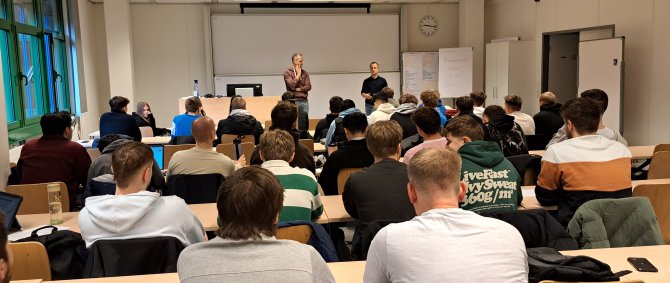 In einem Seminarraum sitzen Studierende und Schüler*innen in Tischreihen, vorne stehen ein Dozent und ein Lehrer und sprechen zur Gruppe.