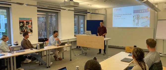 In einem Seminarraum sitzen an zwei Tischreihen Studierende, vorne steht ein junger Mann an einem Schreibtisch mit Laptop, hinter ihm an der Wand eine Präsentationsfolie mit Text: "Gematik - Gastvortrag Hochschule Flensburg"