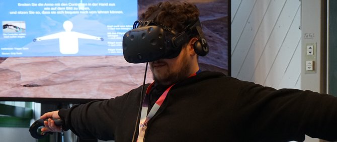 Ein junger Mann trägt eine VR-Brille, hat zwei Controller in den Händen und streckt die Arme zu beiden Seiten aus. Im Hintergrund sieht man einen Bildschirm, auf dem eine virtuelle Umgebung abgebildet ist.