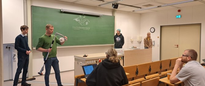 Eine Gruppe von Studierenden steht vorne in einem Vorlesungssaal und hält eine Präsentation. Einer der Studierenden hält ein elekrtisches Gerät mit Teleskoparm in der Hand, an der Tafel hinter ihnen ist ein Logo angezeichnet mit Schriftzug "Dachrinnen-Held"