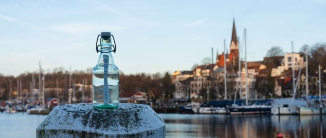 Der Flensburger Hafen an einem sonnigen Wintermorgen. Im Hintergrund sieht man Segelschiffe, eine Promenade und einen Kirchturm, im Vordergrund steht eine durchsichtige Bügelverschluss-Flasche auf einem Poller, in ihr ein aufgerollter Zettel.