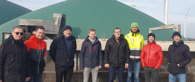 Eine Gruppe von Männern verschiedenen Alters steht vor einer Biogasanlage, im Hintergrund sieht man Windräder, alle lächeln in die Kamera.