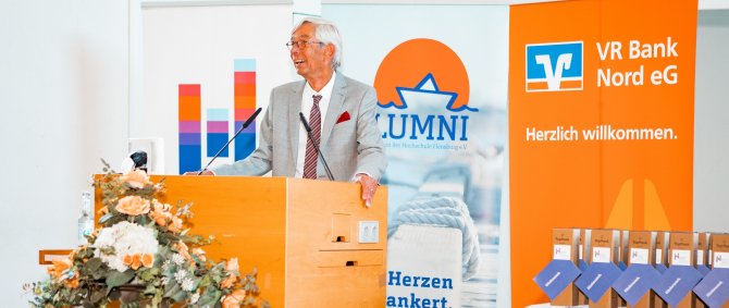 Ein älterer Mann im grauen Anzug steht an einem Rednerpult, vor ihm ein Blumenstrauß, hinter ihm drei Rollups, das mittlere mit dem Logo des Alumni- und Fördervereins der Hochschule Flensburg.