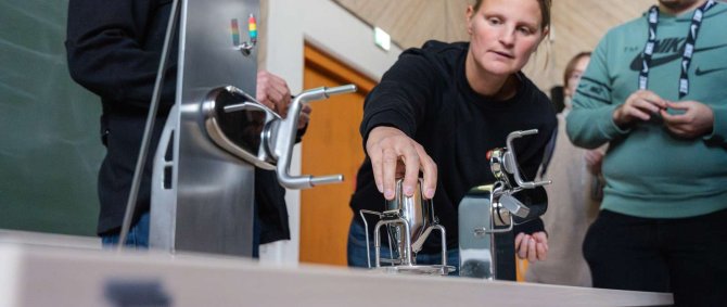 Auf einem Tisch stehen verschiedene Metall-Geräte, eine junge Frau beugt sich über den Tisch und greift danach.