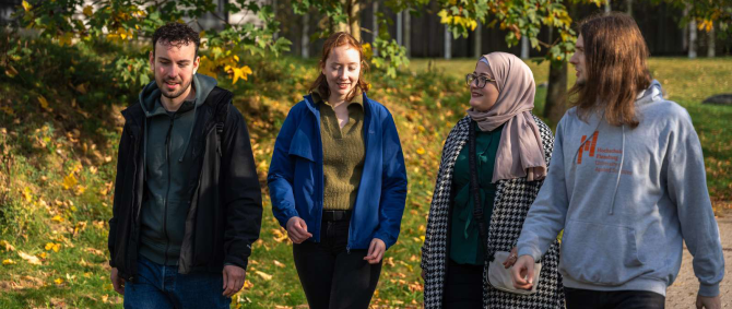 Vier junge Menschen laufen im Sonnenschein über einen Gehweg, im Hintergrund eine grüne Wiese und Bäume. Eine von ihnen trägt ein Kopftuch, ein anderer trägt einen grauen Kapuzenpulli mit dem Logo der Hochschule Flensburg.