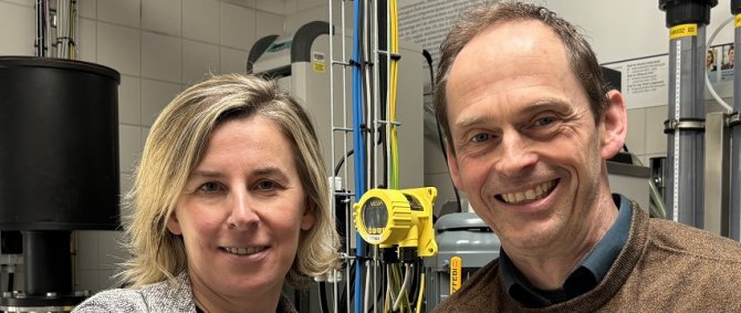 Ein Mann im brauen Pullover und eine Frau im grauen Blazer mit roter Hose stehen in einem Labor an einem Kessel, um sie herum viele Schläuche und Ventile. Beie lächeln freundlich in die Kamera.