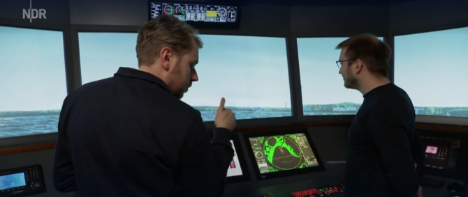 Zwei junge Männer stehen im Schiffbrückensimulator der Hochschule Flensburg, einem Raum, der ausgestattet ist die die Brücke eines großen Schiffes. Aus den Fenstern sieht man eine Projektion des Travemünder Hafens.