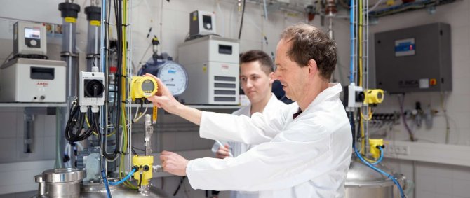 Ein Student und ein Dozent in Laborkitteln stehen in einem Labor an einem Tank, an den viele Schläuche und Ventile angeschlossen sind.